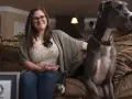 Donna sorride seduta sul divano accanto al cane più grande del mondo, un alano grigio con un collare colorato.