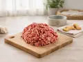 Un mucchio di lonza di maiale per cani macinata su un tagliere di legno, con biscotti a forma di osso e una ciotola per cani sullo sfondo.
