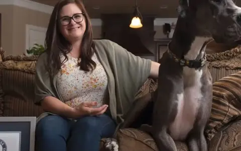 Donna sorride seduta sul divano accanto al cane più grande del mondo, un alano grigio con un collare colorato.